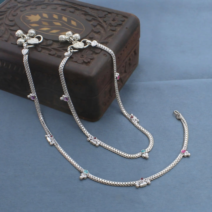 A pair of silver anklets with colorful beads is spread across a light blue surface. The jewelry rests next to an ornate, dark brown wooden box.