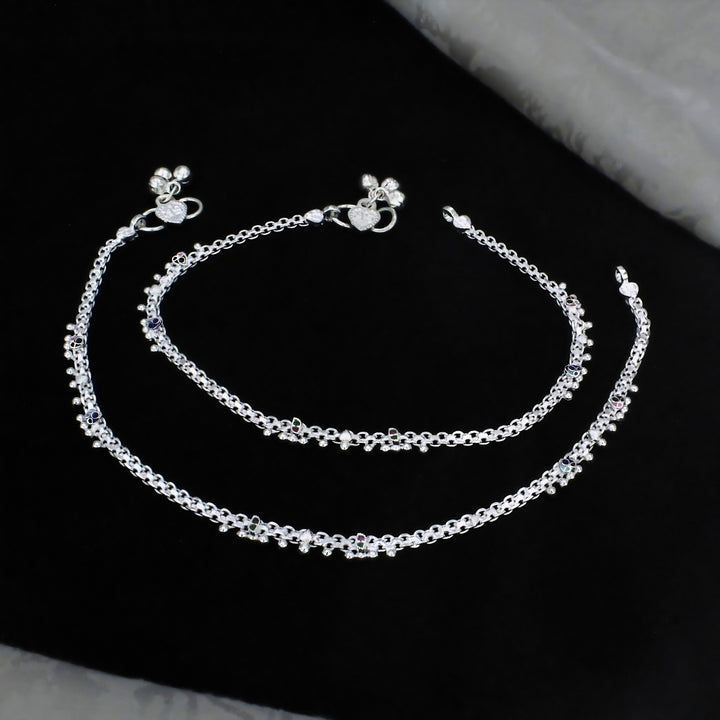 A pair of silver anklets with tiny charms and colored stones, laid out on a dark black fabric background.