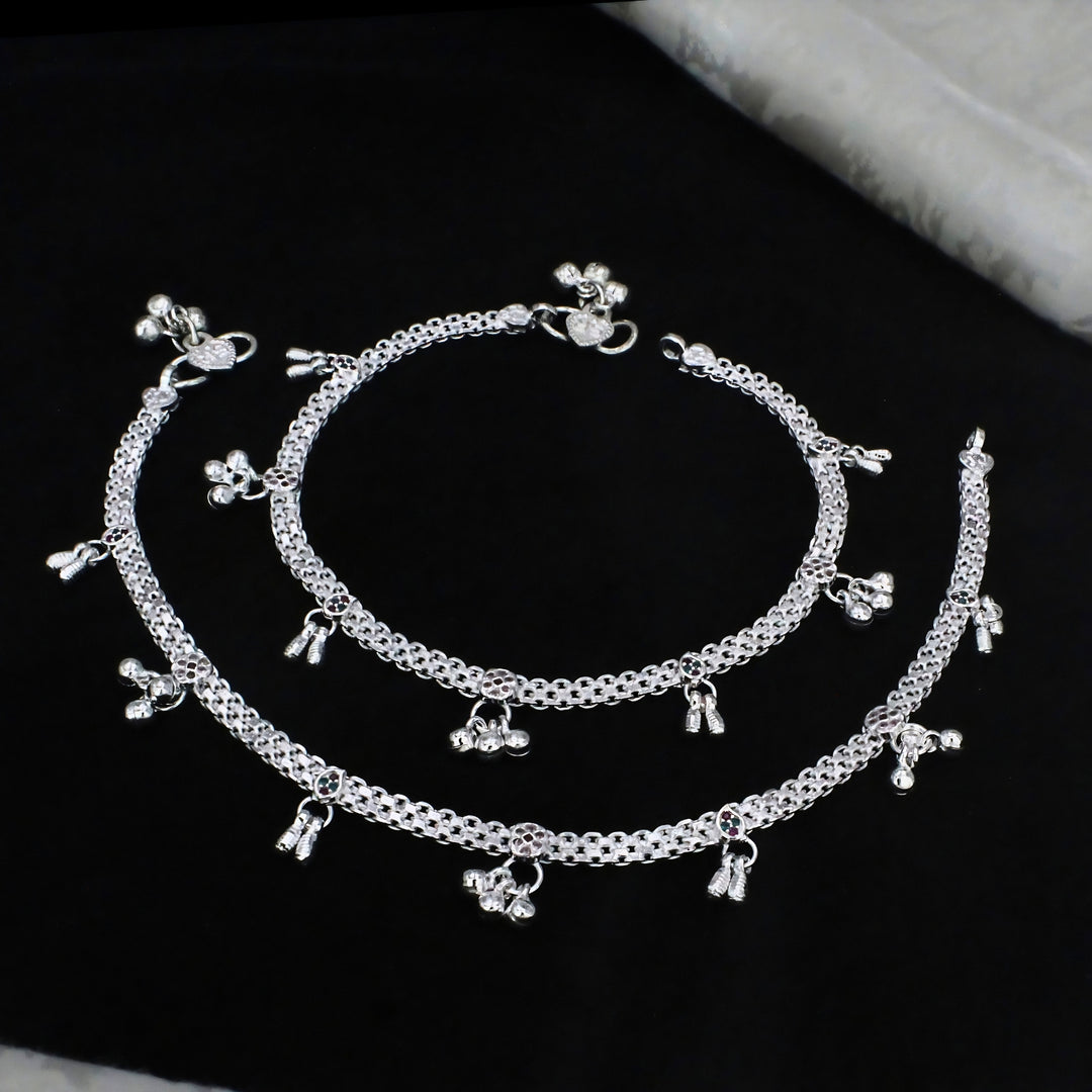 A pair of ornate sterling silver anklets featuring intricate mesh chains, floral charms, and jingle bells, elegantly arranged on a textured black background.
