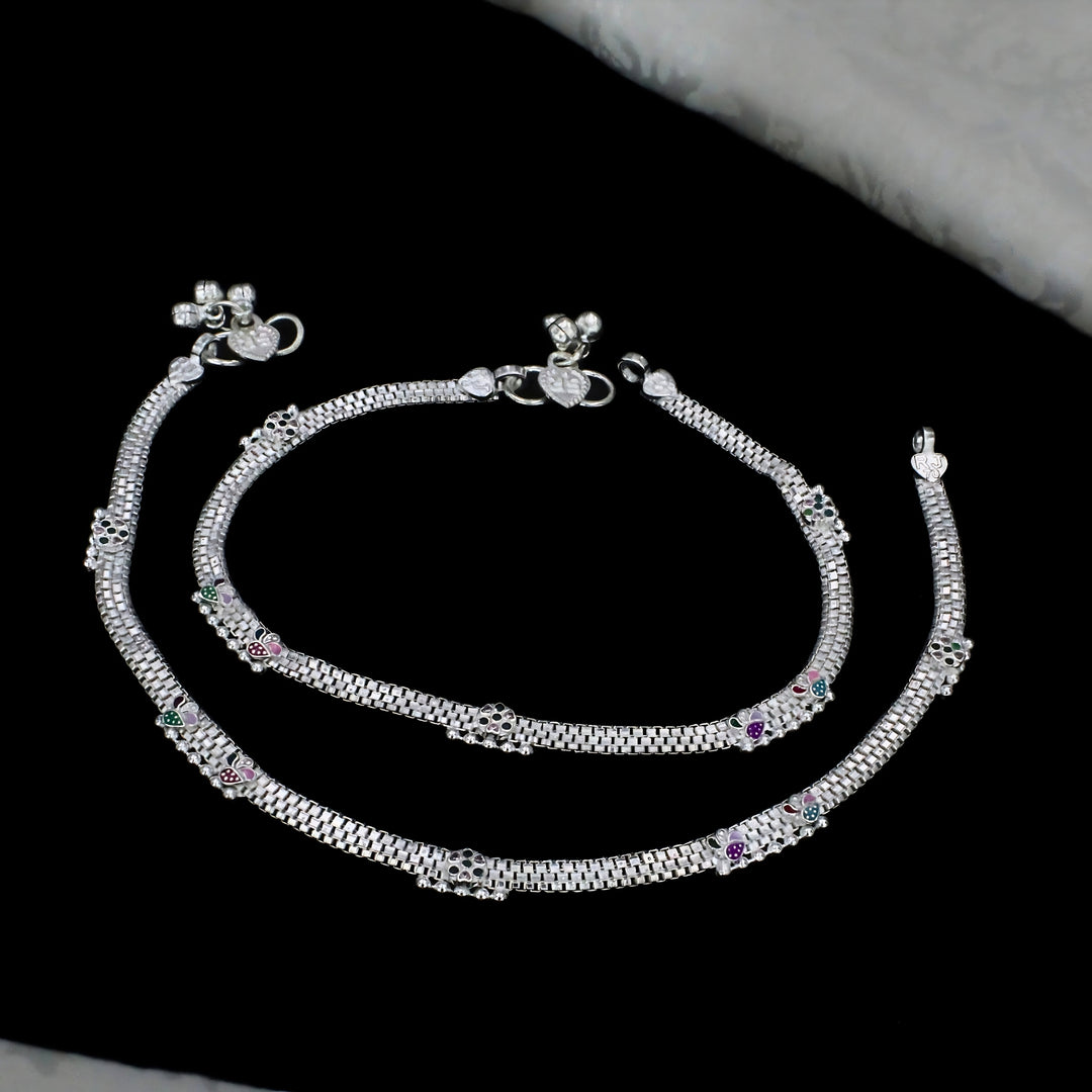 A pair of ornate silver payal anklets featuring mesh chain patterns, colorful enamel accents, and decorative jingle bells clasps, elegantly arranged on a black background.