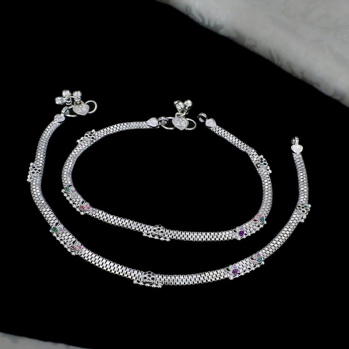 A pair of ornate silver payal anklets featuring mesh chain patterns, colorful enamel accents, and decorative jingle bells clasps, elegantly arranged on a black background.