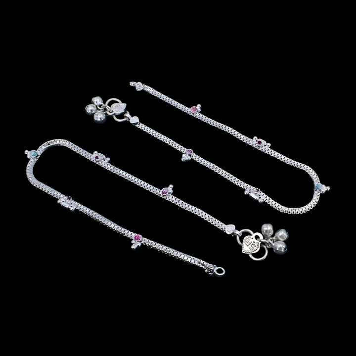 A pair of silver anklets with small, colorful stones and hanging bells, displayed diagonally against a solid black background.