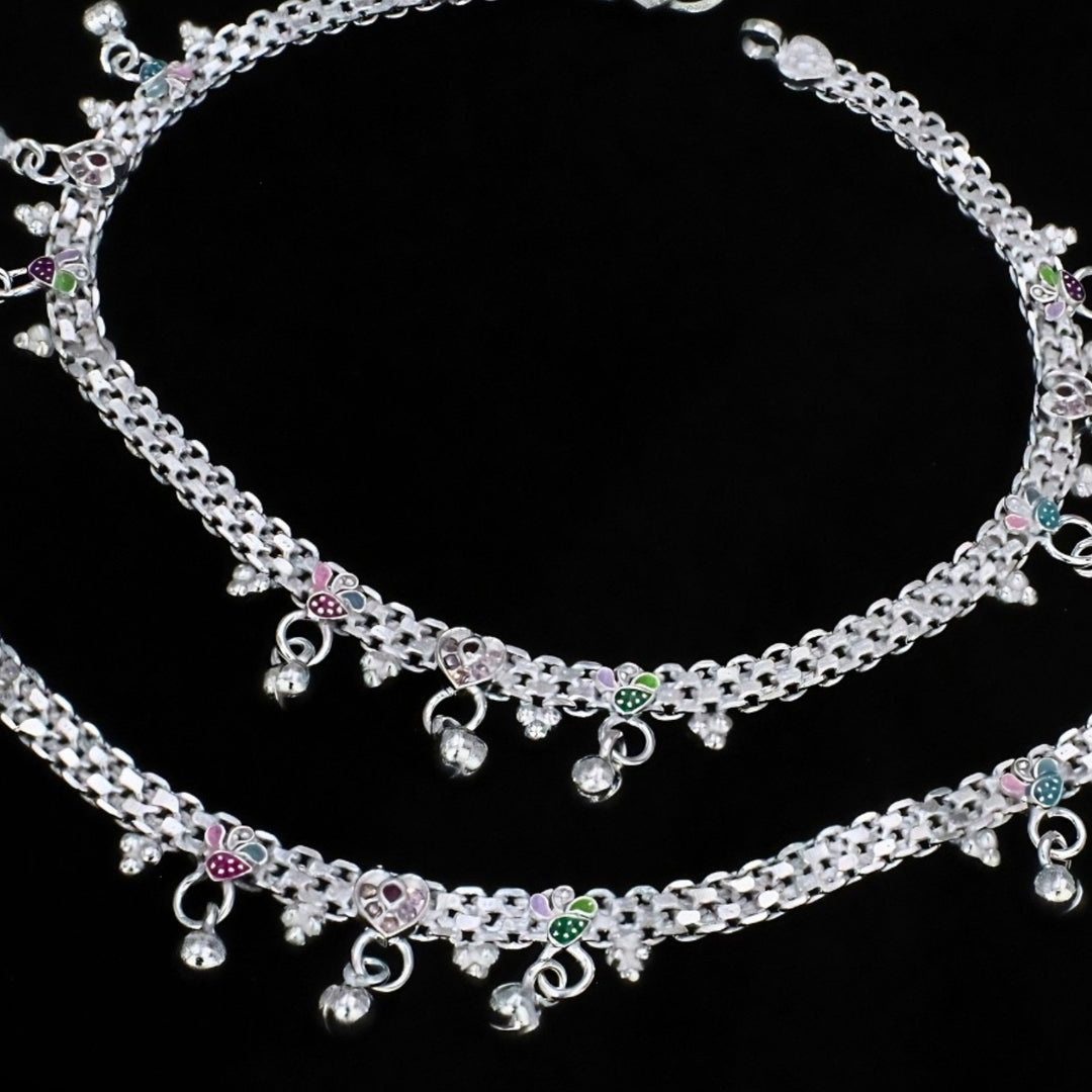 Two silver anklets with colorful leaf charms and small hanging bells are displayed against a black background.