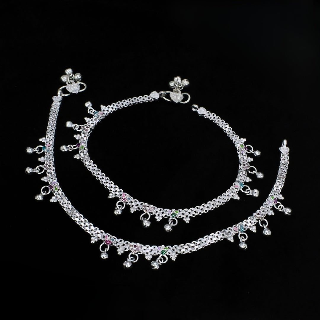 An overhead shot of two silver anklets with small bells and silver beads, arranged in a curve against a solid black background.