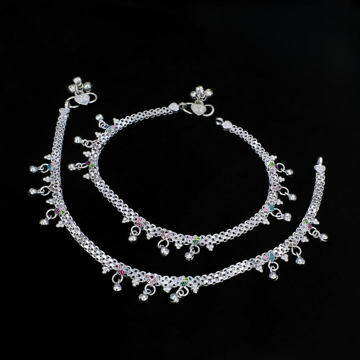 An overhead shot of two silver anklets with small bells and silver beads, arranged in a curve against a solid black background.