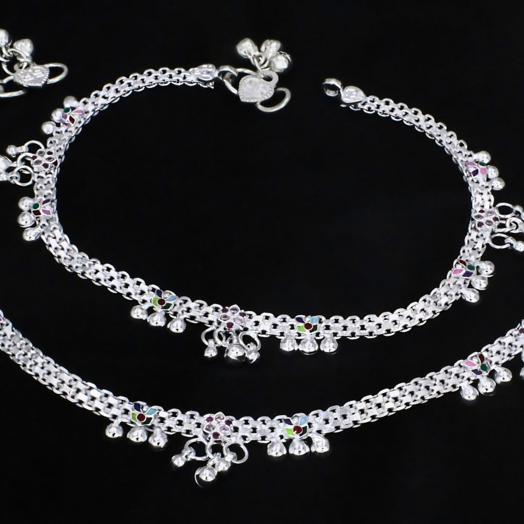 Aerial view of a pair of intricate silver payal anklets with woven chain design, colorful enamel accents, and delicate jingle bells, elegantly arranged on a matte black background.