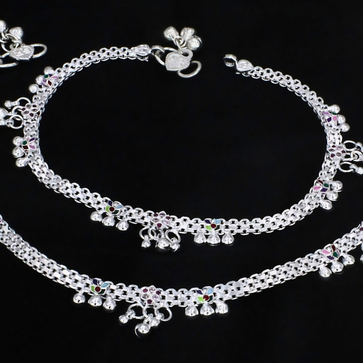 Aerial view of a pair of intricate silver payal anklets with woven chain design, colorful enamel accents, and delicate jingle bells, elegantly arranged on a matte black background.