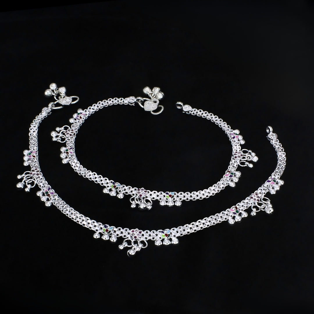 A pair of intricately designed silver payal anklets featuring detailed chainwork, colorful enamel accents, and dangling jingle bells, elegantly displayed against a solid black background.