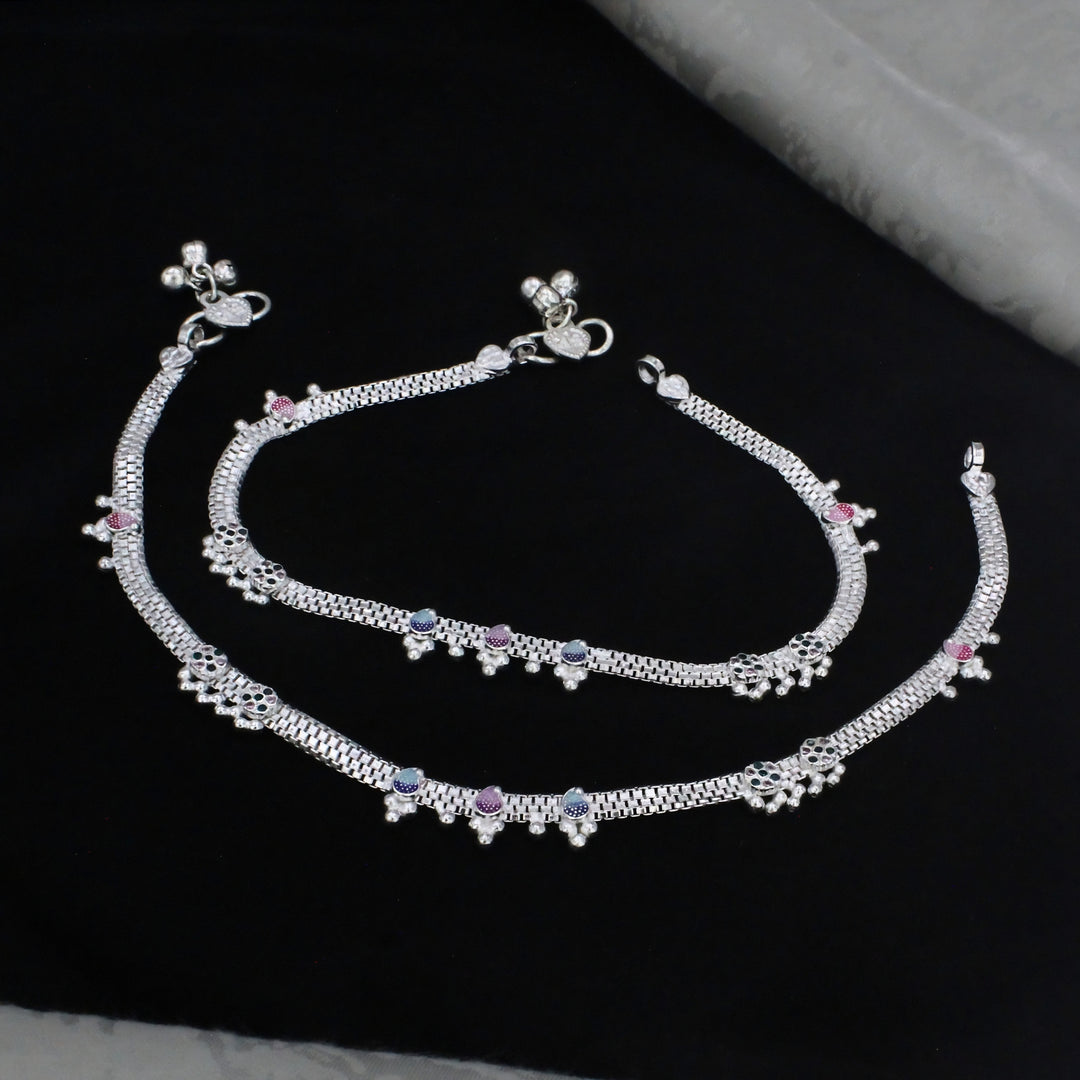 A pair of intricately designed silver anklets featuring textured chain patterns, colorful pink and blue accents, and delicate dangling jingles bells, elegantly displayed on a dark black background.