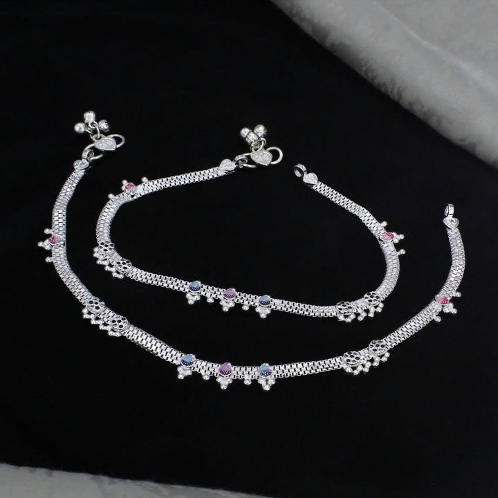 A pair of intricately designed silver anklets featuring textured chain patterns, colorful pink and blue accents, and delicate dangling jingles bells, elegantly displayed on a dark black background.