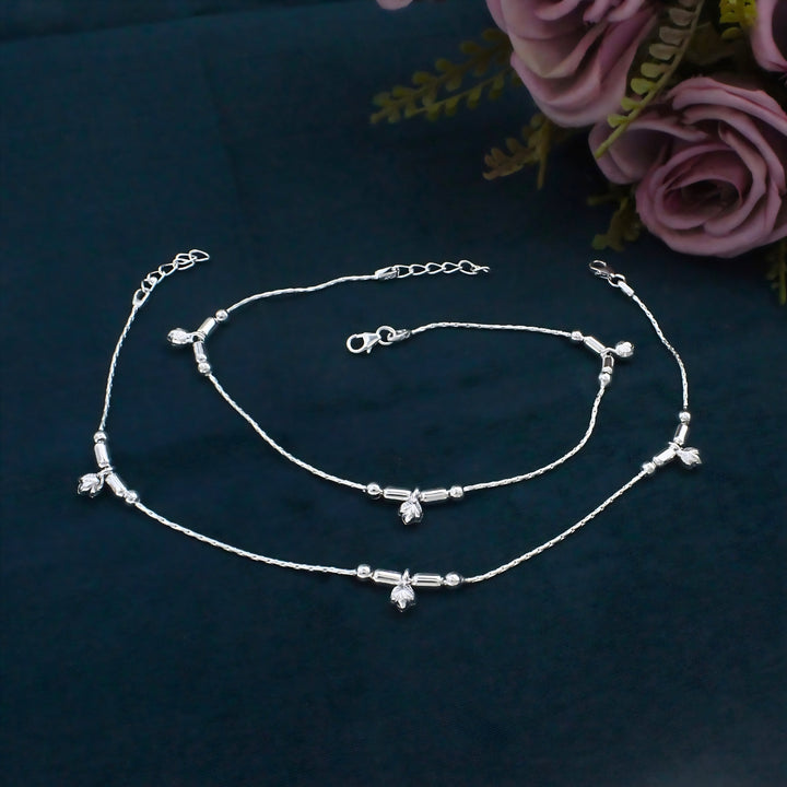 A pair of delicate silver anklets with tiny charms rests on a dark blue fabric surface next to pink artificial roses.
