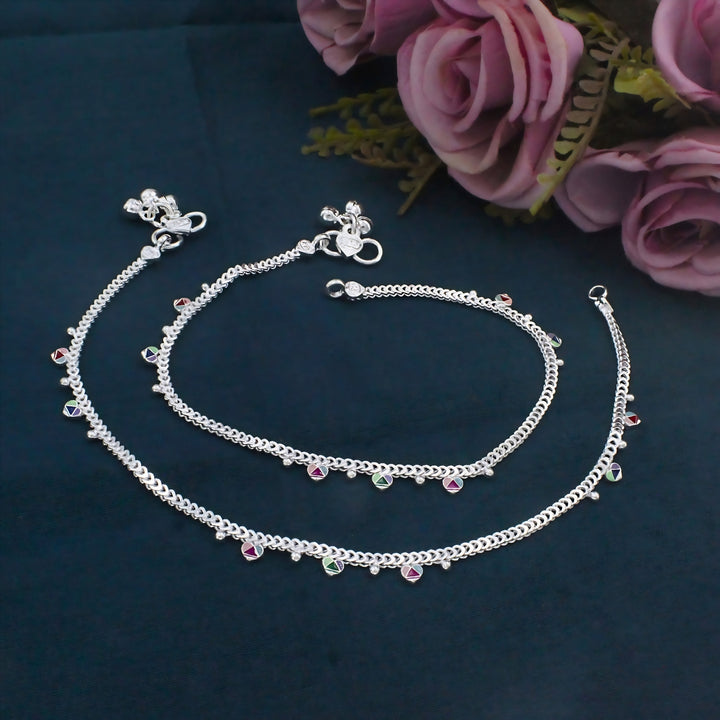 Real silver anklets pair with colorful small charms are laid out on a dark blue surface next to pink roses.